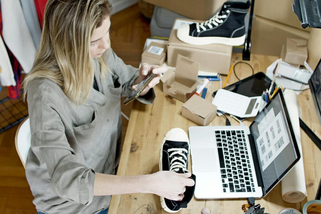 pexels-photo-7289723-7289723 Woman photographing shoes for online sale in home workspace with laptop and packages.